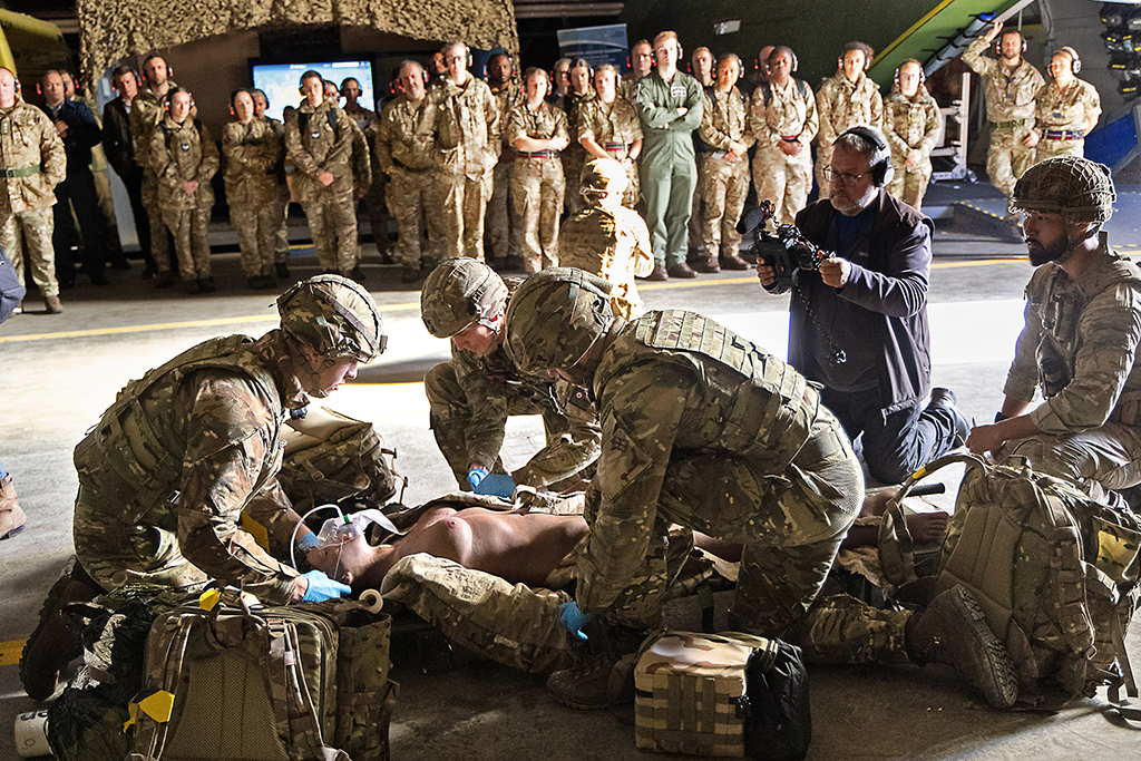 TMW medical personnel, in camouflage uniforms and helmets, perform emergency medical training on a simulation mannequin lying on a stretcher. A cameraman films the exercise in the foreground, while a large group of service members stands in rows watching from the background inside a hangar or transport aircraft.